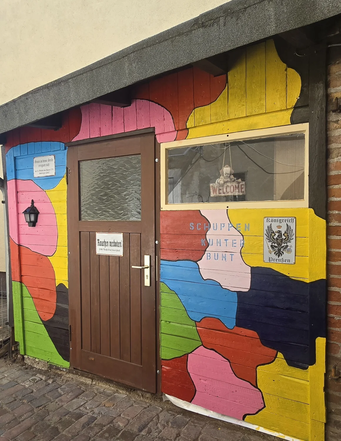 Kunterbunt bemalter Holzschuppen mit der Aufschrift 'Schuppen Kunterbunt'.