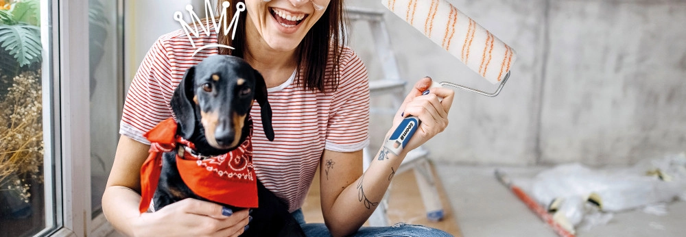 Lachende Person mit Farbspritzern und Hund hält einen Farbroller.