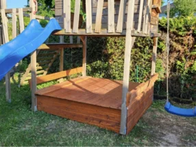 Ein hölzerner Sandkasten unter einem Spielhaus mit Rutsche.