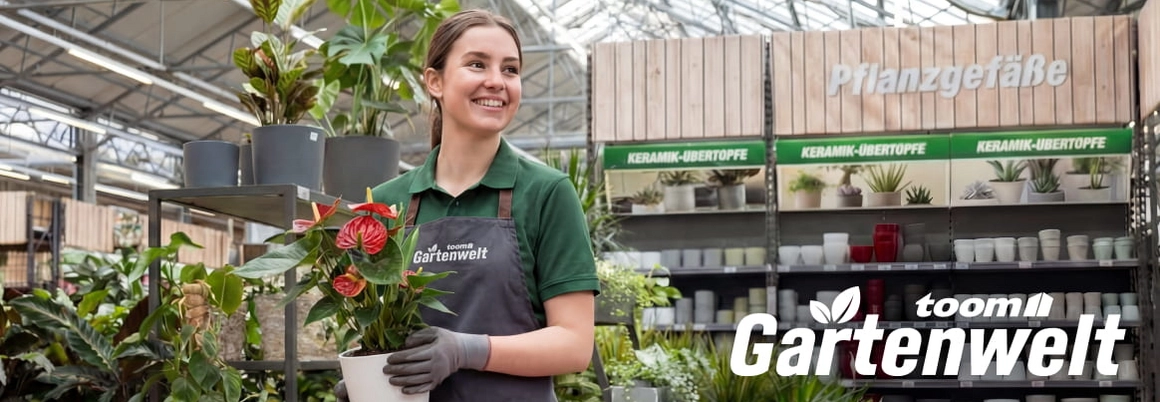 Eine lächelnde Person in einem Gartencenter hält eine blühende Pflanze in einem Topf.