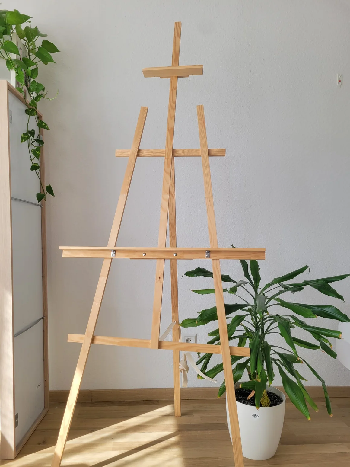 Eine leere Staffelei aus Holz steht in einem Raum neben zwei Zimmerpflanzen.