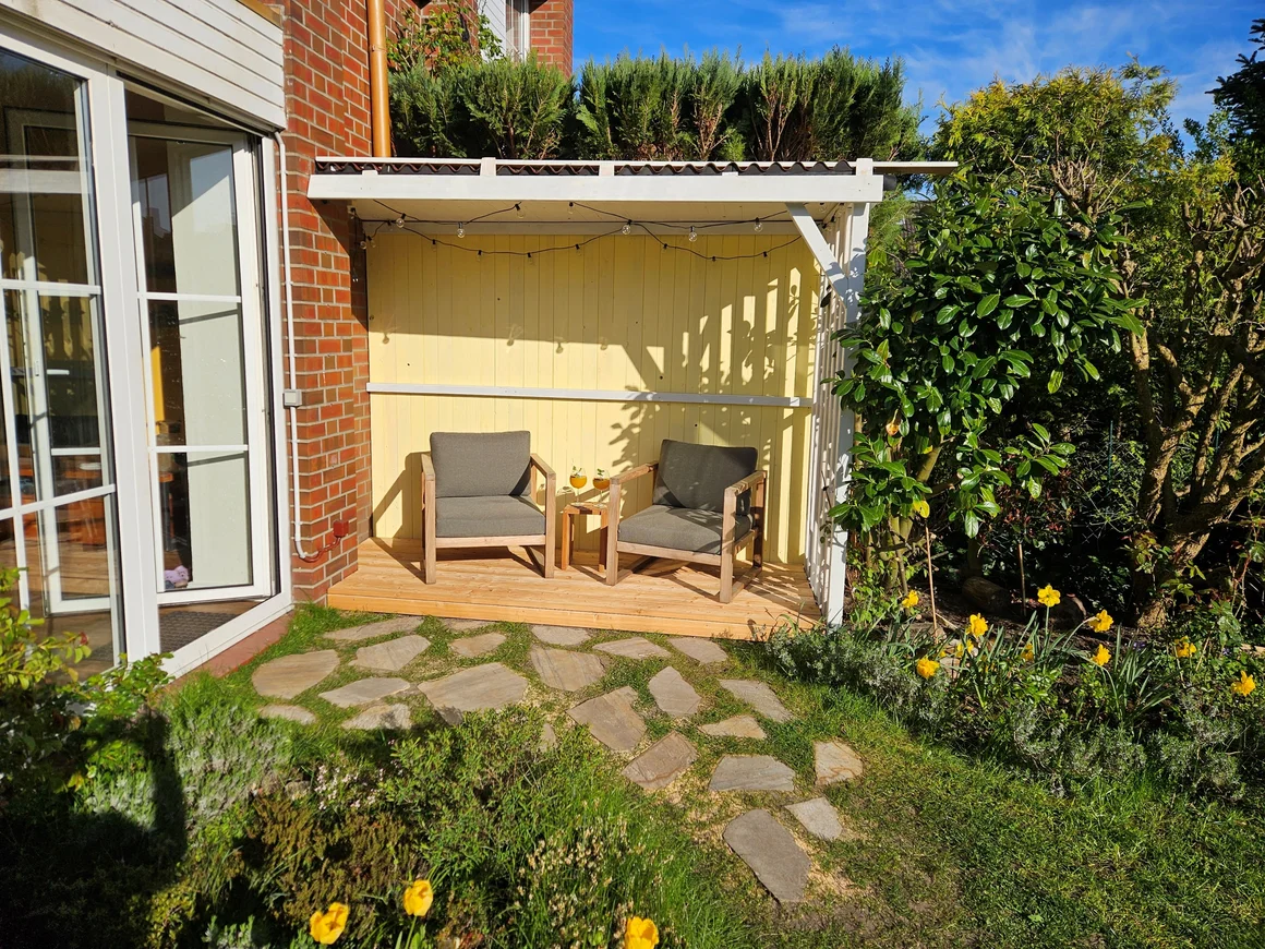 Überdachte Sitzecke mit zwei Sesseln in einem sonnigen Garten.