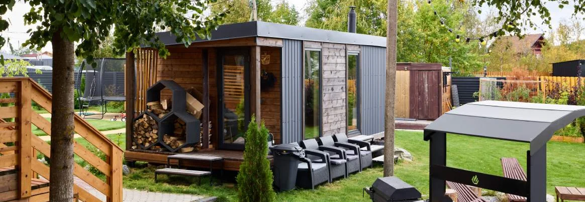 Moderne Gartensauna aus Holz mit Sitzgelegenheiten und Grill im Grünen.