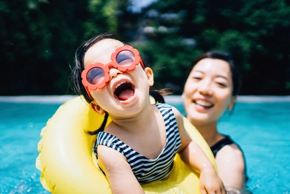 Ein lachendes Kind mit Sonnenbrille und Schwimmring in einem Pool.