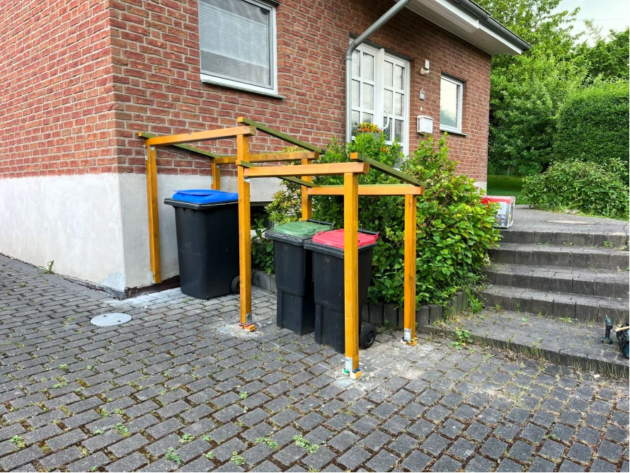 Drei Mülltonnen unter einem Holzgerüst.