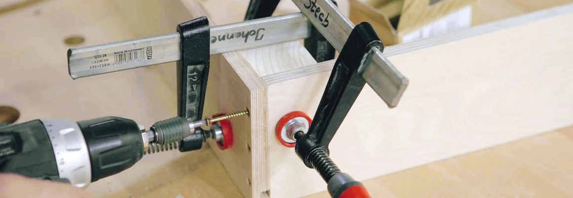 Zwei Holzbretter werden mit einer Schraube und einem Akkuschrauber verbunden und mit einer Schraubzwinge fixiert.