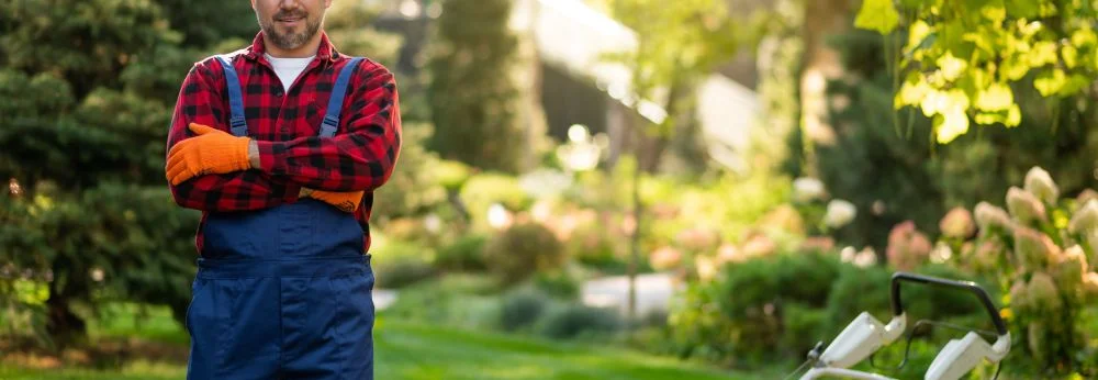 Person in Gärtnerkleidung mit Rasenmäher in einem Garten.
