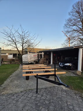 Ein Anhängergestell mit Holzlatten steht auf einer Einfahrt vor einer Garage.