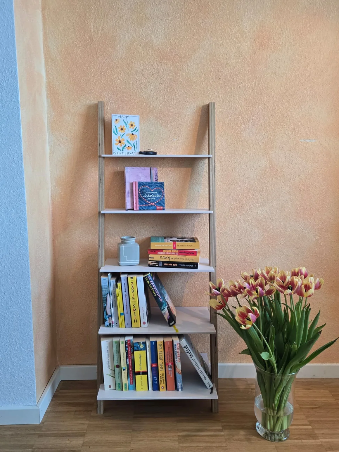 Ein Leiterregal mit Büchern steht neben einer Vase mit bunten Tulpen.