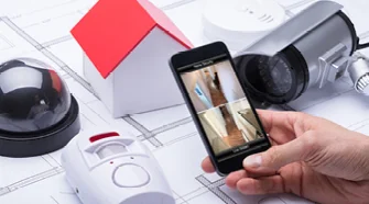 Ein Rauchmelder liegt auf einem Bauplan, daneben ein Schraubenzieher und ein Hausmodell.