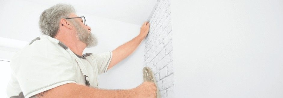 Ein Handwerker bringt die erste Tapetenbahn auf eine Wand auf.