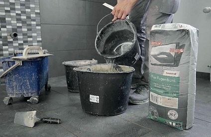 Fugenmörtel für Bodenfliesen vorbereiten Ein Handwerker gibt Wasser in einen großen Eimer mit Fugenmörtel.