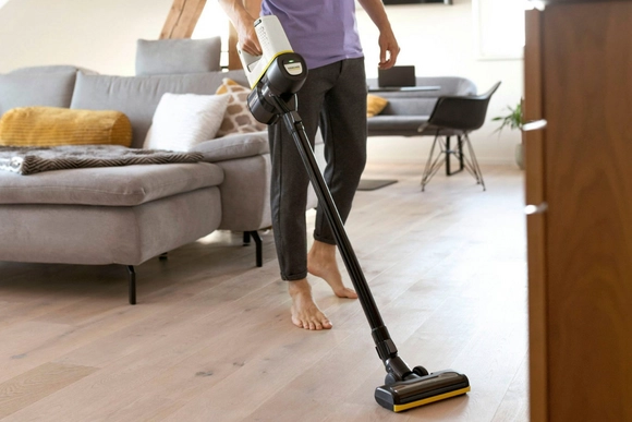 Eine Person saugt barfuß mit einem Akku-Staubsauger einen hellen Holzboden.