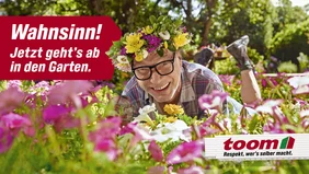 Eine lachende Person mit einem Blumenkranz auf dem Kopf liegt bäuchlings in einem bunten Blumenbeet.