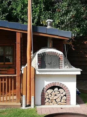 Ein weißer, gemauerter Pizzaofen mit Holzvorrat steht im Garten neben einer Holzhütte.
