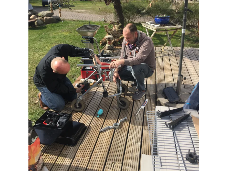 Zwei Personen reparieren eine Gehhilfe mit Werkzeug auf einer Holzterrasse