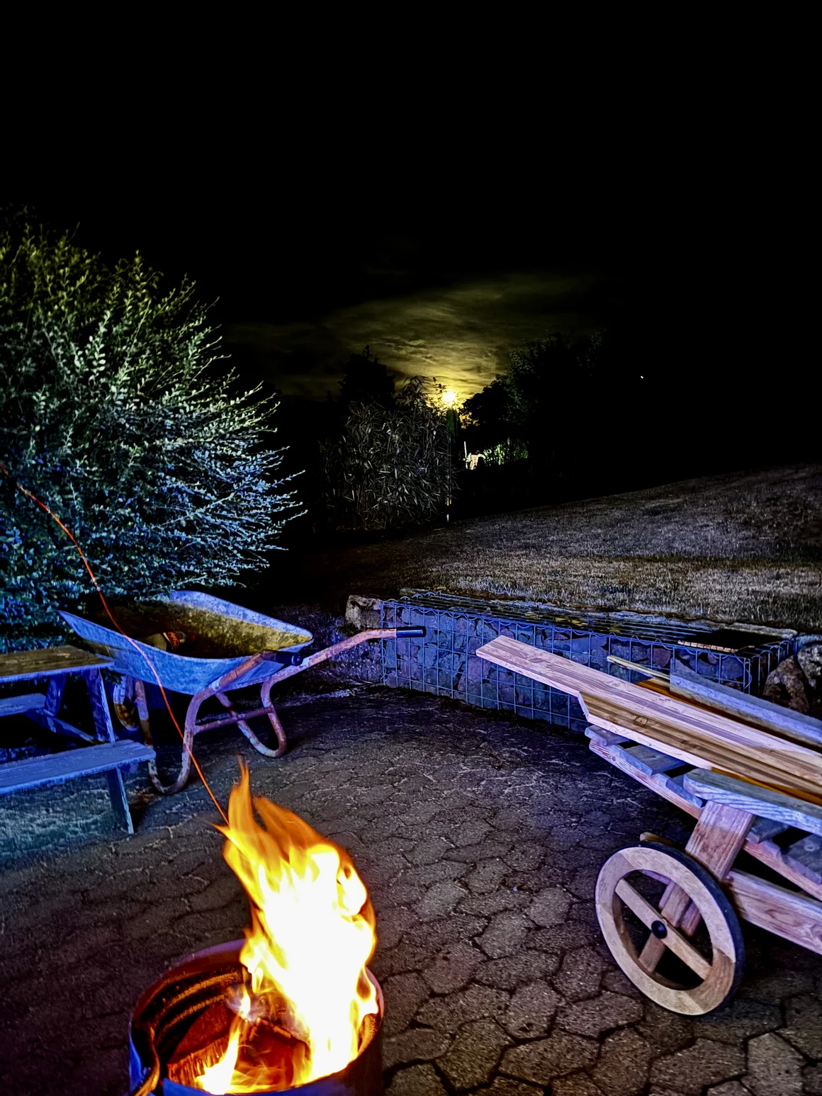 Lagerfeuer im Garten bei Nacht mit Schubkarre und Mondschein.