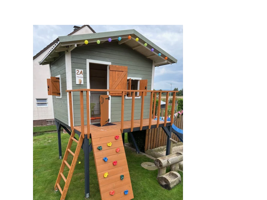 Ein Spielhaus aus Holz auf Stelzen mit einer Rutsche und einer Kletterwand.