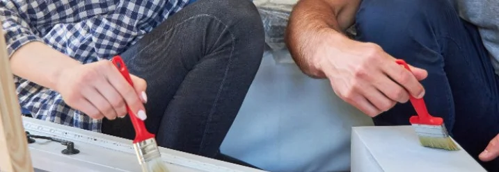 Zwei Personen streichen ein Möbelstück mit Pinseln weiß.