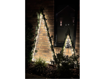 Zwei beleuchtete, hölzerne Weihnachtsbäume vor einem Haus.