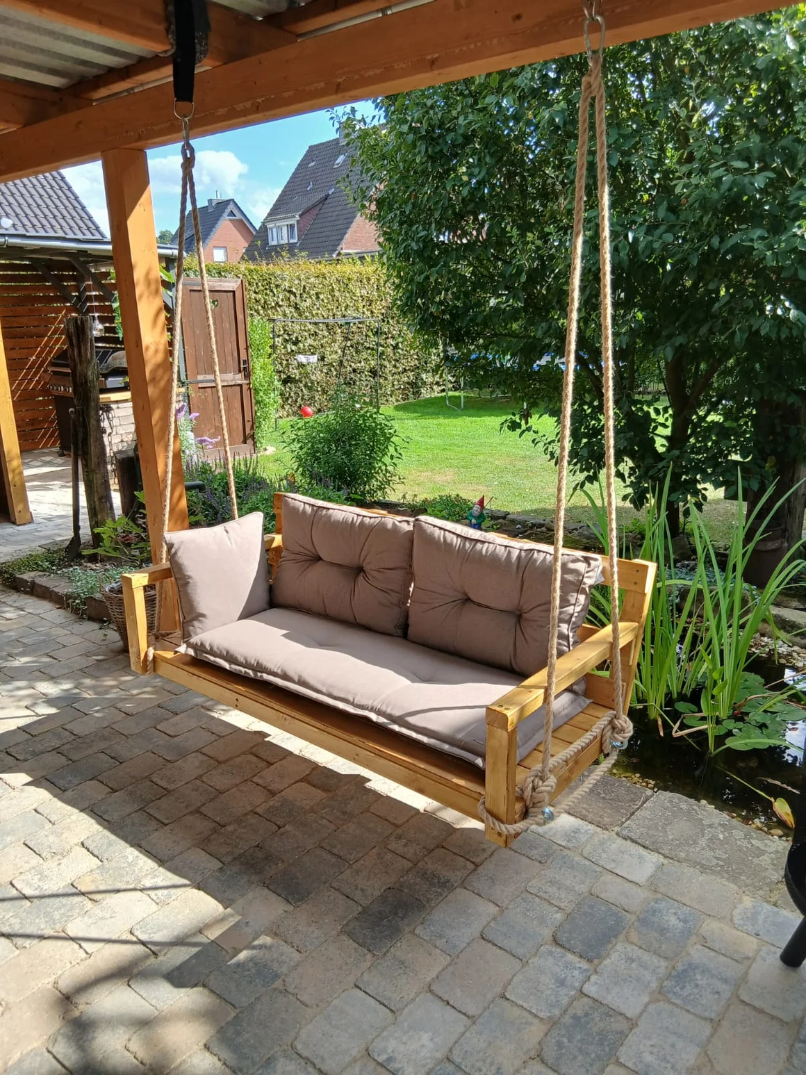 Hölzerne Hollywoodschaukel mit grauen Polstern auf einer Terrasse.