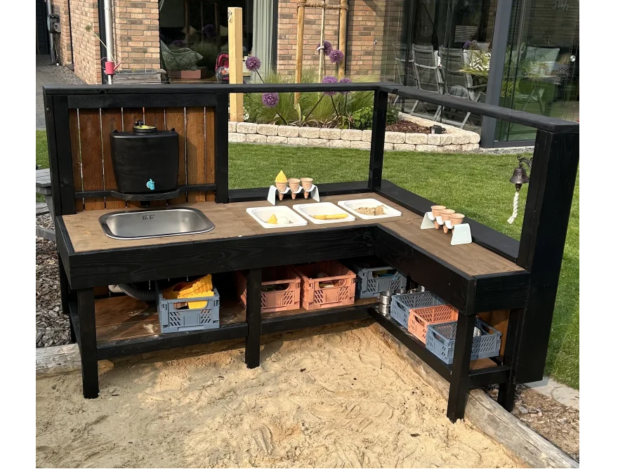 Eine schwarze, L-förmige Matschküche aus Holz steht in einem Sandkasten im Garten.
