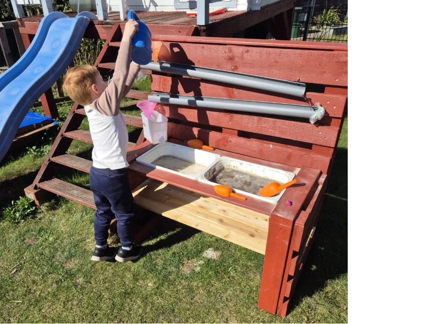 Kind gießt mit Gießkanne Wasser in Spielküche im Freien