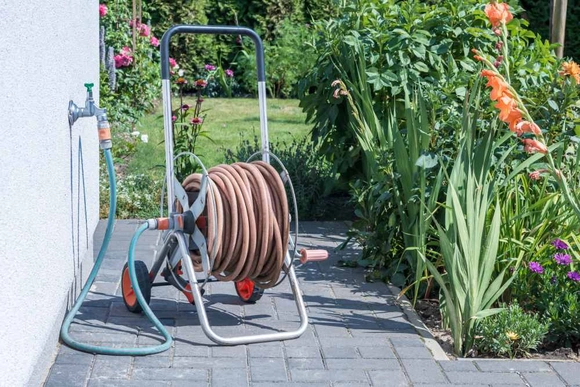 Ein aufgerollter Gartenschlauch auf einem Schlauchwagen steht im Garten an einer Hauswand.