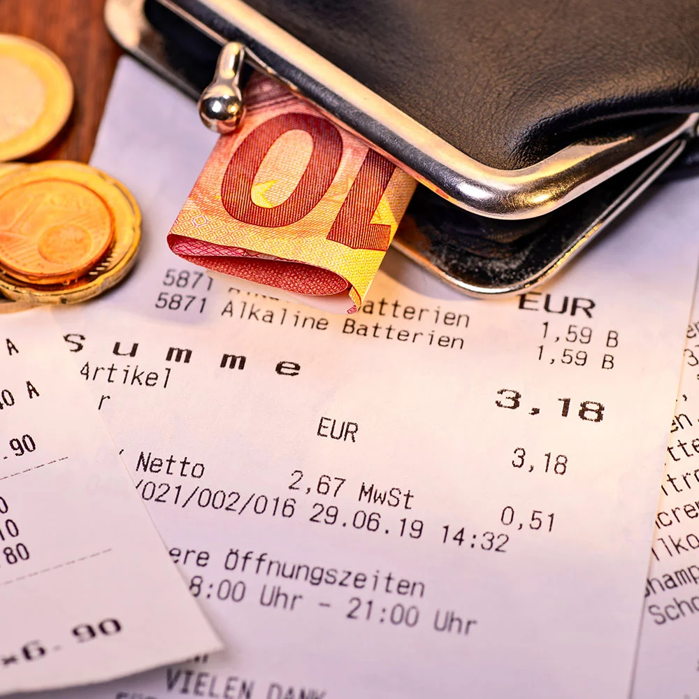Eine schwarze Geldbörse liegt auf einem Kassenbon und es schauen ein Zehn-Euro-Schein sowie drei Zwei-Euro-Münzen heraus.