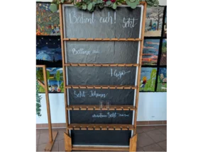 Ein hölzernes Regal mit mehreren Ebenen, in denen Sektgläser aufbewahrt werden können. Die Ebenen haben schwarze Tafeloberflächen, auf denen verschiedene Sektsorten aufgeschrieben sind.