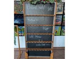 Ein hölzernes Regal mit mehreren Ebenen, in denen Sektgläser aufbewahrt werden können. Die Ebenen haben schwarze Tafeloberflächen, auf denen verschiedene Sektsorten aufgeschrieben sind.