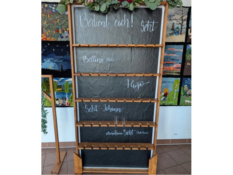 Ein hölzernes Regal mit mehreren Ebenen, in denen Sektgläser aufbewahrt werden können. Die Ebenen haben schwarze Tafeloberflächen, auf denen verschiedene Sektsorten aufgeschrieben sind.