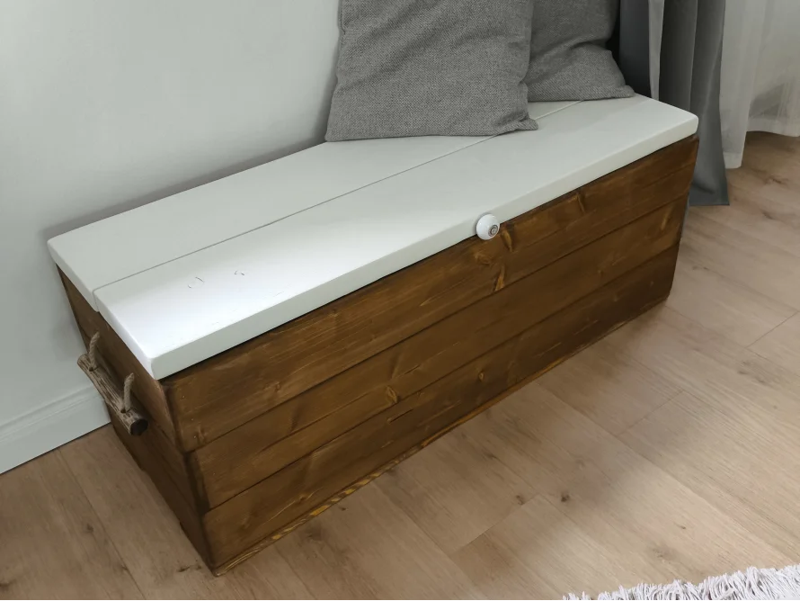 Eine braune Holztruhe mit weißem Deckel und Kissen dient als Sitzgelegenheit.