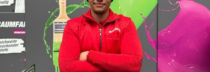 Eine lächelnde Person in roter Jacke vor einer Wand mit Pinsel-Motiv.
