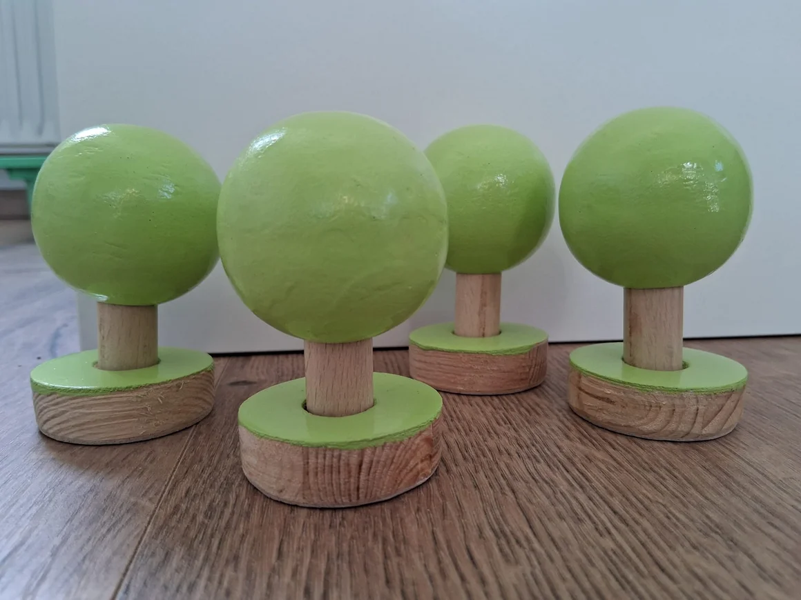 Vier stilisierte Bäume aus Holz mit grünen Kronen auf einem Holzboden.