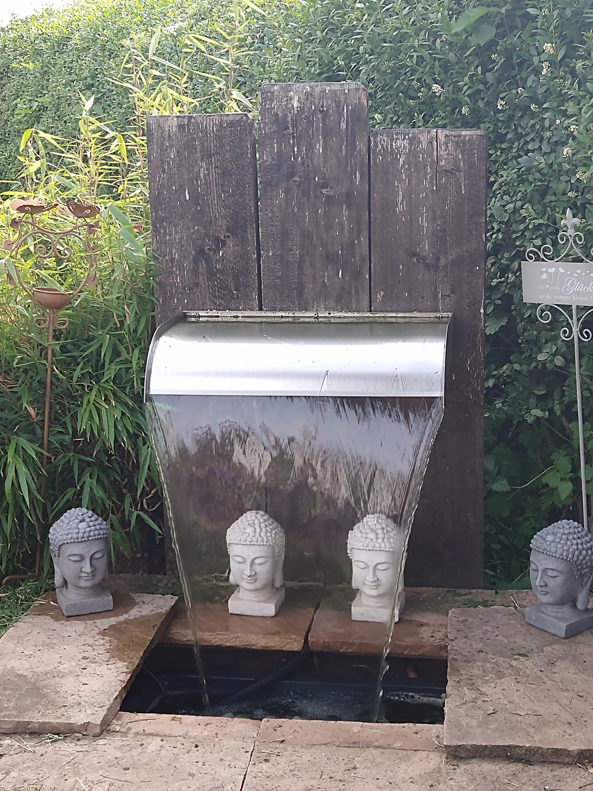 Ein Gartenbrunnen als Wasserfall mit Buddha-Kopf-Dekorationen und einer Holzwand.
