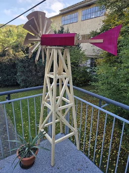 Ein großes, selbstgebautes Windrad aus Holz mit pinkem Dach auf einem Balkon.