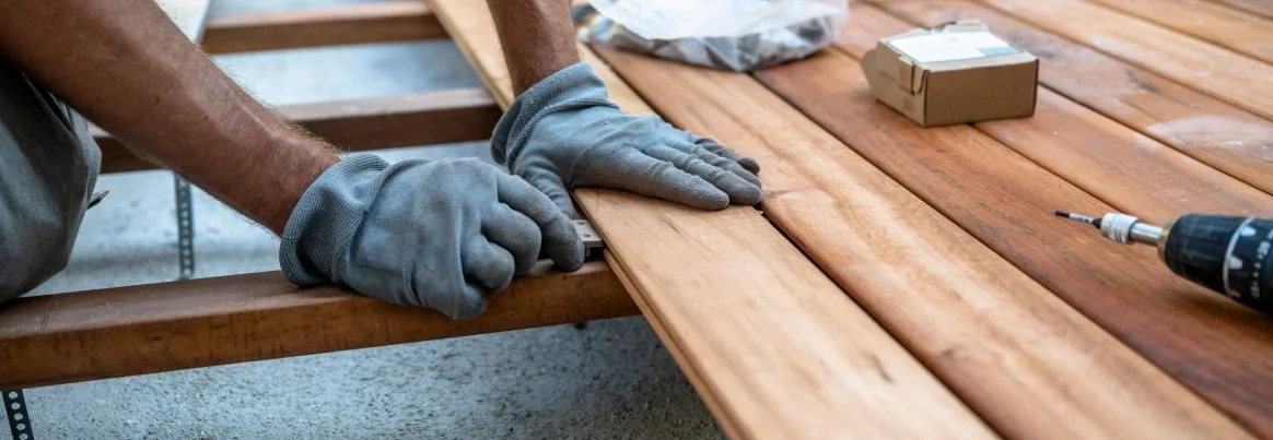 Eine Person verlegt mit Handschuhen Holzdielen auf einer Terrasse.