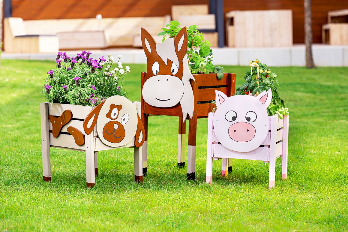 Drei Blumenkästen in Tierform stehen bepflanzt auf einer Wiese.