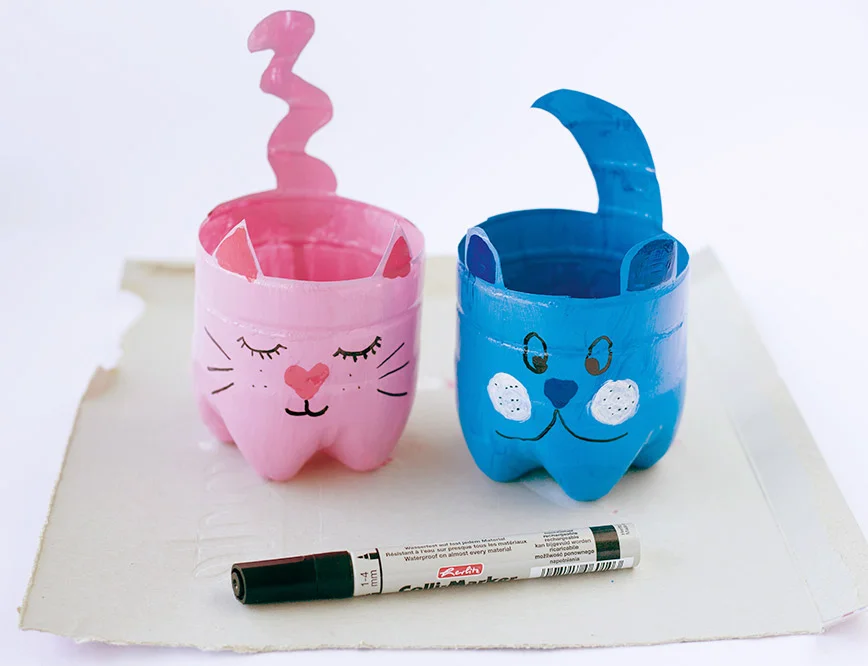 Zwei aus Plastikflaschen gebastelte Katzenköpfe mit Filzstift bemalt