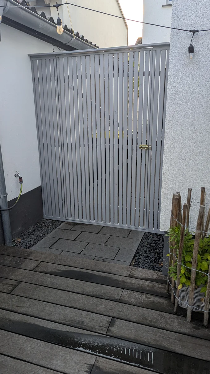 Ein graues Gartentor aus vertikalen Holzlatten neben einer Holzterrasse.