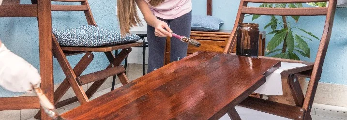 Ein Kind lasiert mit einem Pinsel einen Holztisch auf einem Balkon.