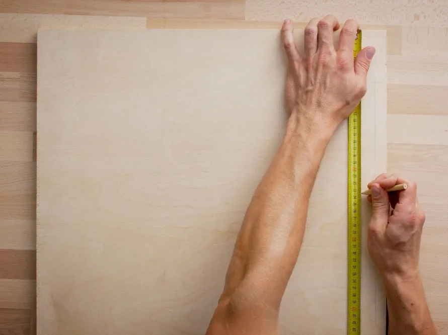 Eine Person misst eine Sperrholzplatte mit einem Zollstock und markiert sie mit einem Bleistift.