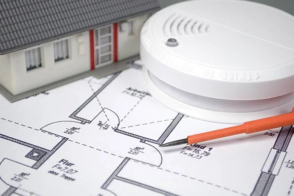 Ein Rauchmelder liegt auf einem Grundriss neben einem orangen Schraubendreher und einem Hausmodell.