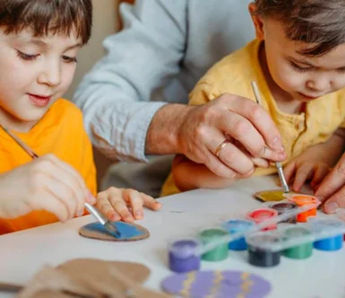 Zwei Kinder und eine erwachsene Person bemalen Pappfiguren mit Pinseln.