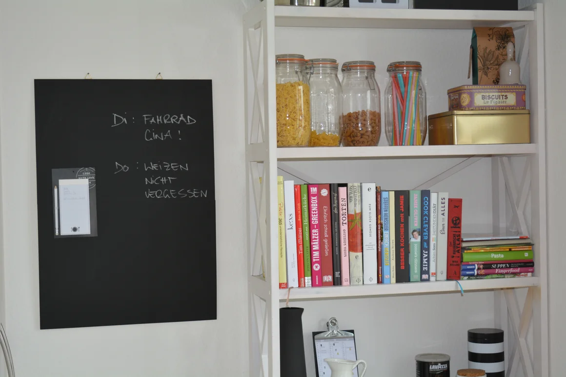 Ein weißes Regal mit Kochbüchern und einer schwarzen Magnettafel