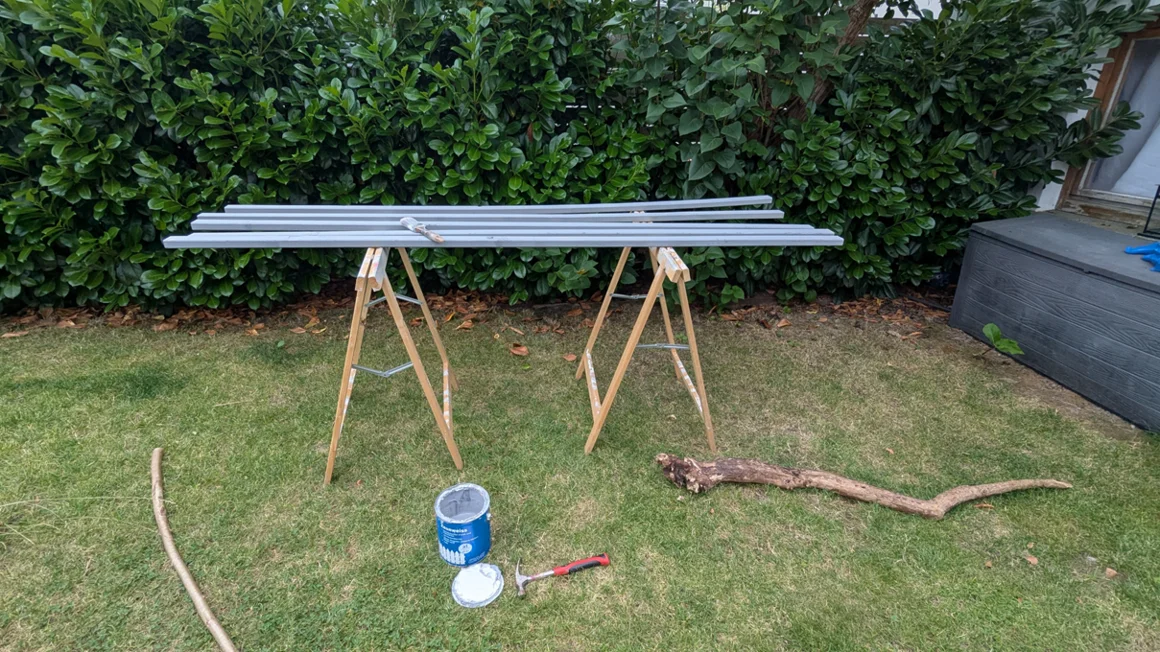 Grau gestrichene Latten liegen auf Böcken im Garten, daneben Farbe und Hammer.
