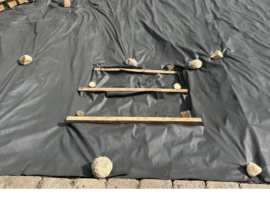 Schwarze Folie mit drei Holzlatten und Steinen