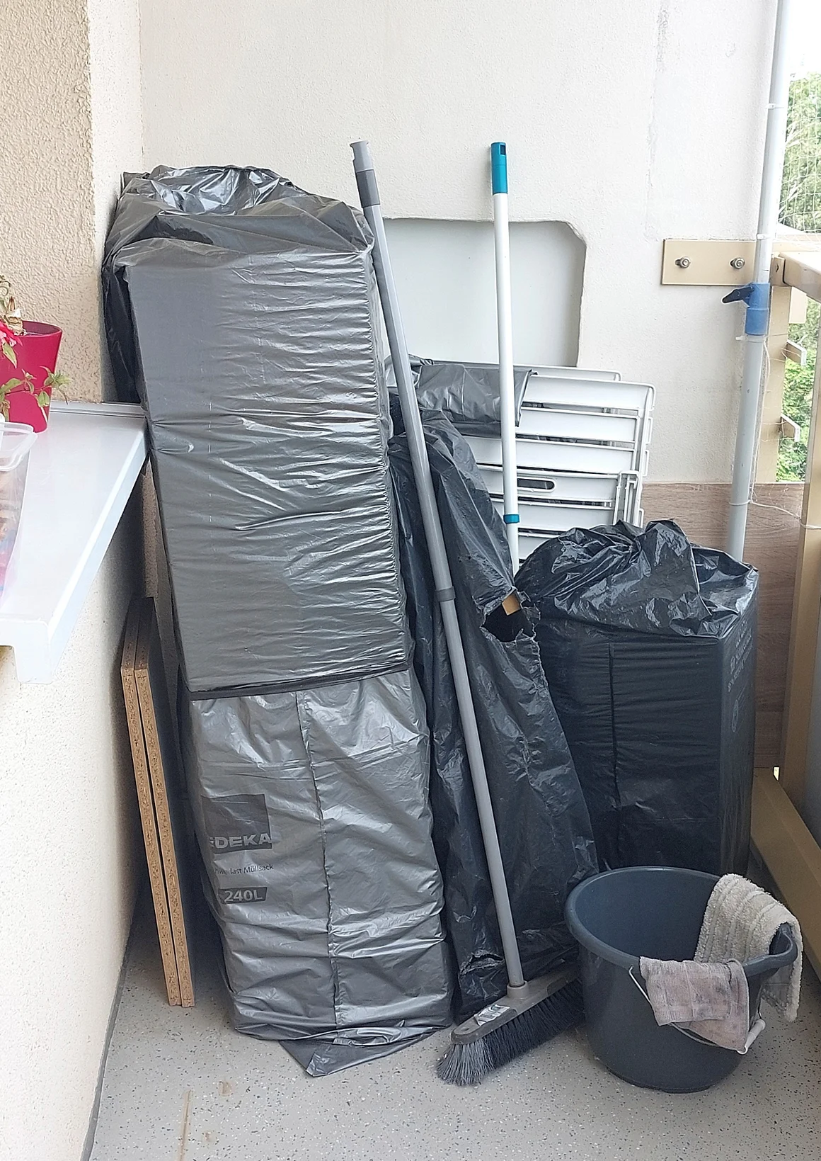 Eine unaufgeräumte Balkonecke mit Besen, Eimer und verpackten Gegenständen.