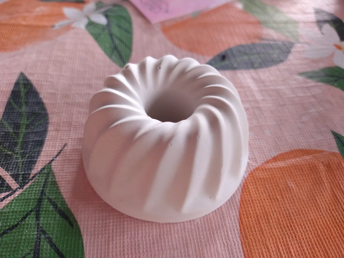 Ein weißer, geriffelter Dekogegenstand in Gugelhupfform liegt auf einer gemusterten Tischdecke.